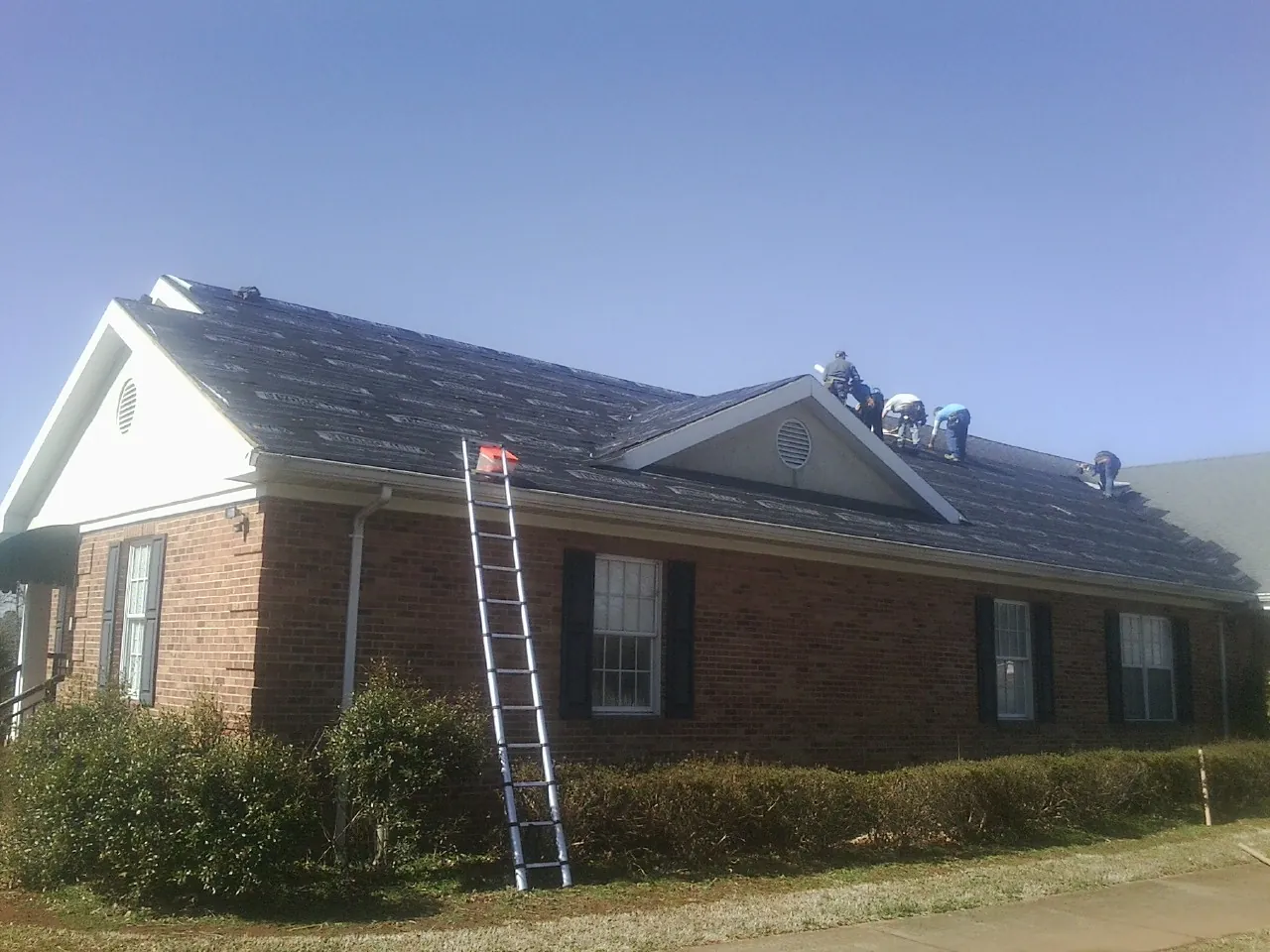 Crew tearing shingles off a brick ranch roof with a ladder set — Emergency Roof Repair work by our crew in West Sand Lake