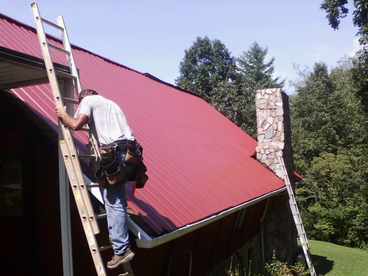 Roofer on a ladder beside a red metal roof and stone chimney — Soffit & Fascia Repair by Roofing Brothers in West Sand Lake